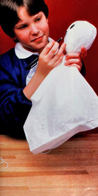 a boy in 1980s-style clothing draws a face on a balloon-and-tissue paper ghost